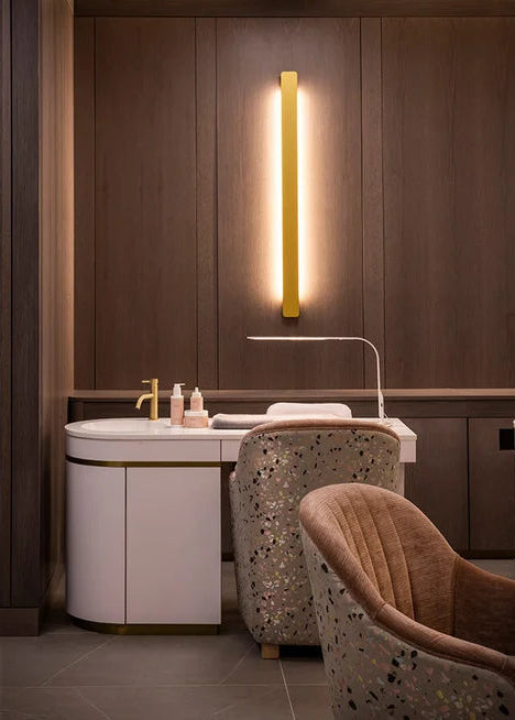 A modern spa or bathroom vanity area with a white sink cabinet, gold faucet, illuminated vertical wall sconce, patterned chairs, and dark wood paneling.