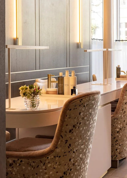 A modern vanity desk setup in a spa or salon interior, featuring a curved white countertop with gold and beige toiletry bottles, a small vase of yellow flowers, and two upholstered chairs with a terrazzo pattern in beige, brown, and black.