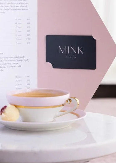 A pink and white Mink Berlin menu card stands on a small round white table next to a white teacup with a gold rim and a small pastry, in a softly lit indoor setting.