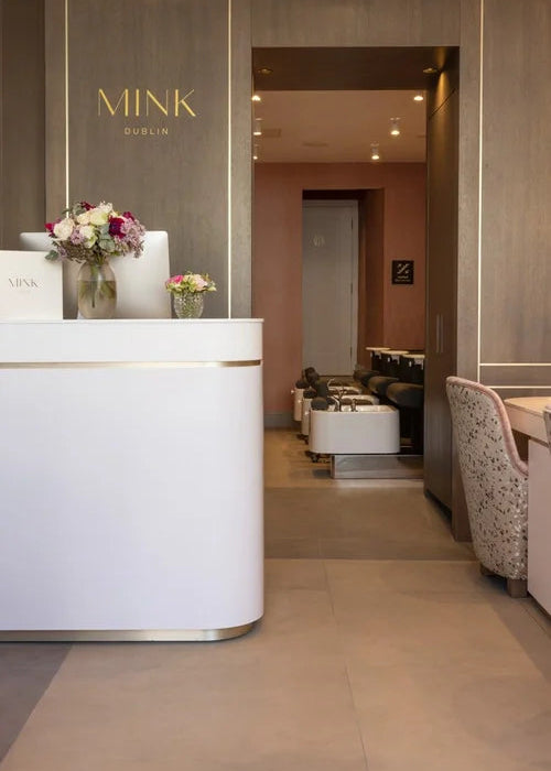 A modern spa or salon reception area with a white curved counter, gold MINK DUBLIN branding on a wood-paneled wall, and a softly lit interior seating area in the background.