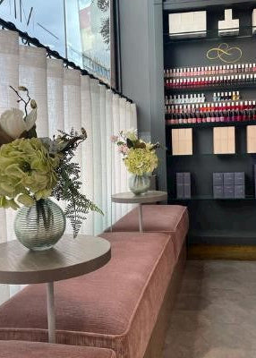 A modern nail salon interior with a long blush pink upholstered bench, small round tables with flower arrangements, and a dark display wall filled with nail polish bottles and product boxes.