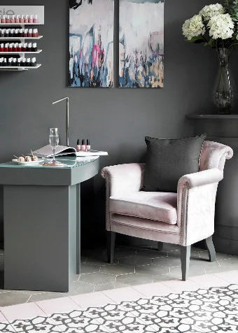 A styled interior scene with a pale blush pink velvet armchair, a dark gray vanity desk, abstract wall art, and a vase of white flowers against a dark gray wall.